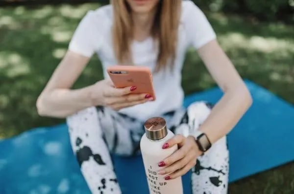 woman on phone sitting yoga mat holding water bottle